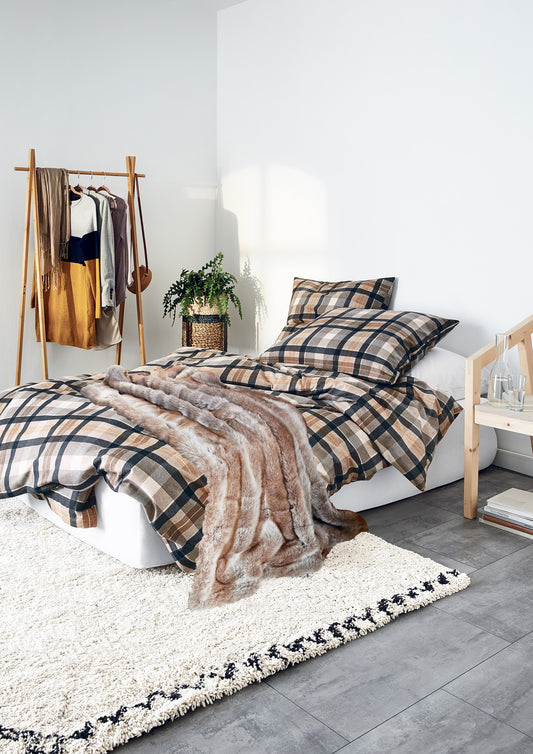 Auf einem Bett liegen mehrere Kopfkissen und eine Bettdecke bezogen mit Edel-Feinbiber Bettwaesche aus 100 Prozent Baumwolle mit Muster aus Quadraten in der Farbe Braun. Modell Koala Natur.