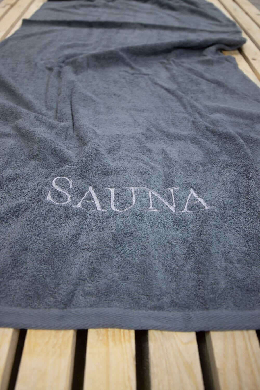 Auf einer Bank liegt ein Saunatuch mit aufgesticktem Text SAUNA von p.k. aus 100 Prozent Baumwolle in der Groesse 70x200cm. Farbe Anthrazit. Modell BW 500 Anthrazit.