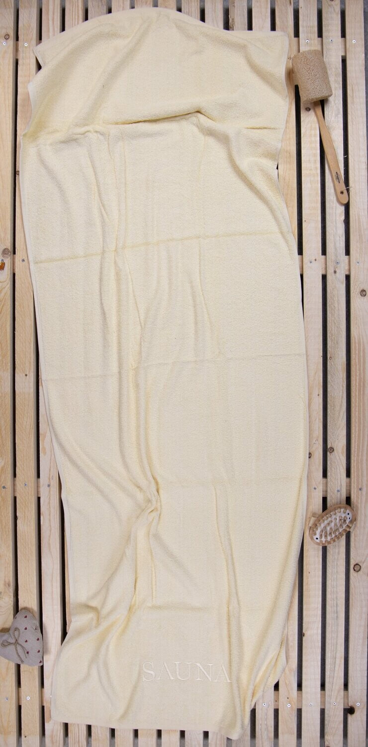 Auf einer Holzbankank liegt ein Saunatuch mit aufgesticktem Text SAUNA von p.k. aus 100 Prozent Baumwolle in der Groesse 70x200cm. Farbe Beige. Modell Creme.
