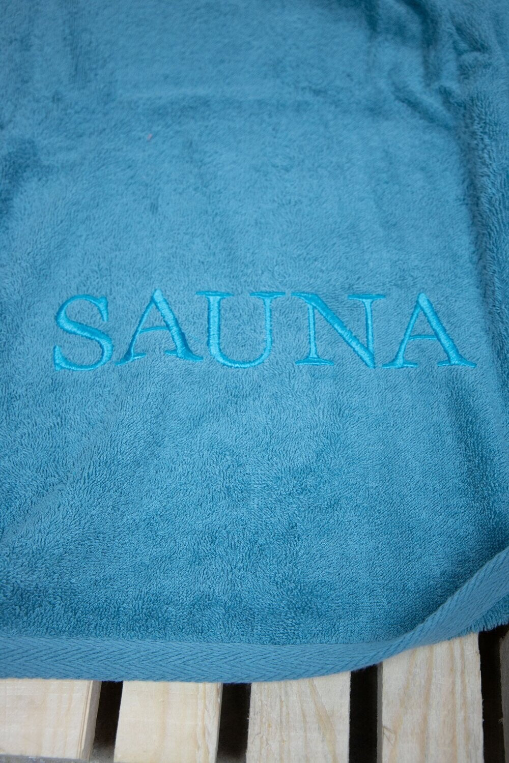Auf einer Bank liegt ein Saunatuch mit aufgesticktem Text SAUNA von p.k. aus 100 Prozent Baumwolle in der Groesse 70x200cm. Farbe Petrol . Modell Petrol