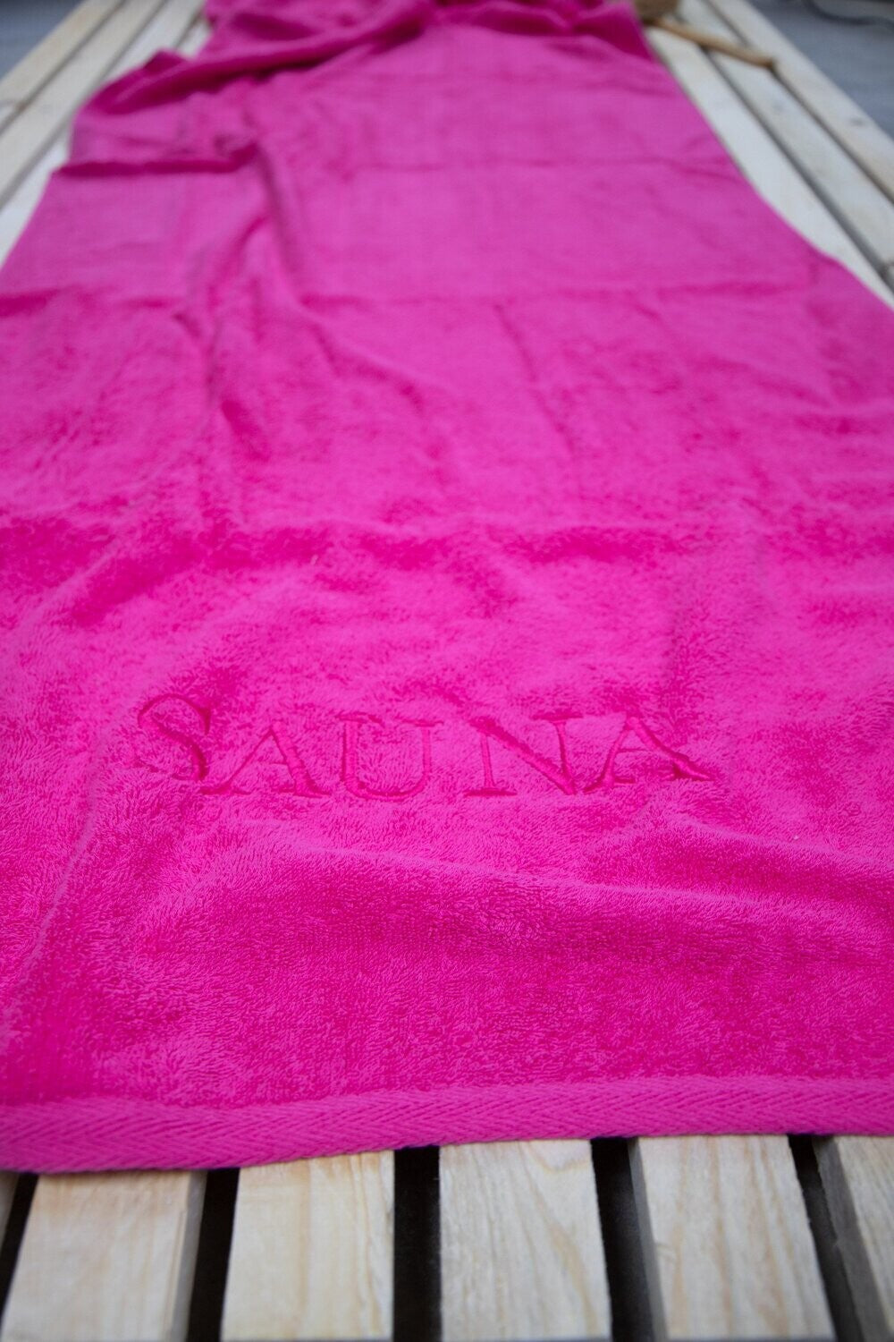 Auf einer Bank liegt ein Saunatuch mit aufgesticktem Text SAUNA von p.k. aus 100 Prozent Baumwolle in der Groesse 70x200cm. Farbe Pink. Modell Pink.