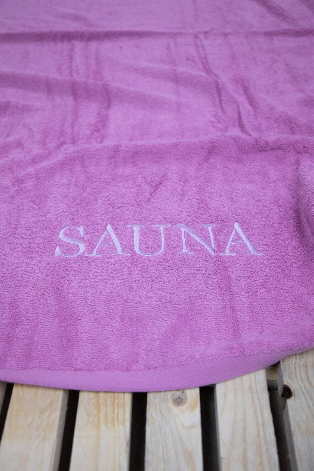 Auf einer Bank liegt ein Saunatuch mit aufgesticktem Text SAUNA von p.k. aus 100 Prozent Baumwolle in der Groesse 70x200cm. Farbe Rosa. Modell Rosenholz.