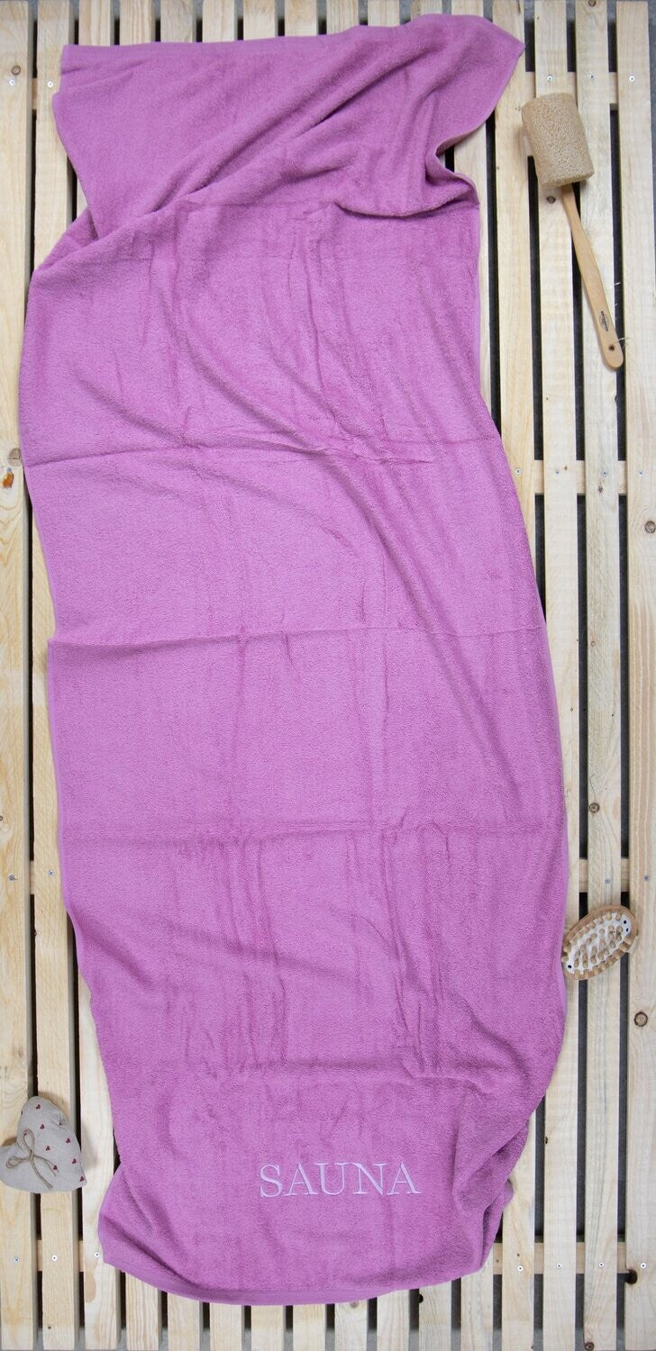 Auf einer Holzbankank liegt ein Saunatuch mit aufgesticktem Text SAUNA von p.k. aus 100 Prozent Baumwolle in der Groesse 70x200cm. Farbe Rosa. Modell Rosenholz.