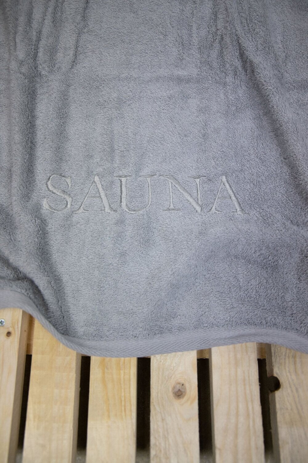 Auf einer Bank liegt ein Saunatuch mit aufgesticktem Text SAUNA von p.k. aus 100 Prozent Baumwolle in der Groesse 70x200cm. Farbe Silber. Modell Silber. 