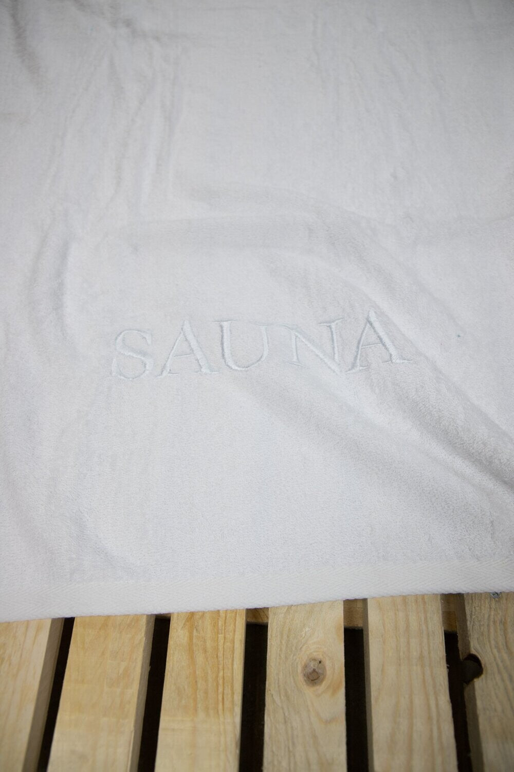 Auf einer Bank liegt ein Saunatuch mit aufgesticktem Text SAUNA von p.k. aus 100 Prozent Baumwolle in der Groesse 70x200cm. Farbe Weiss. Modell Weiss.