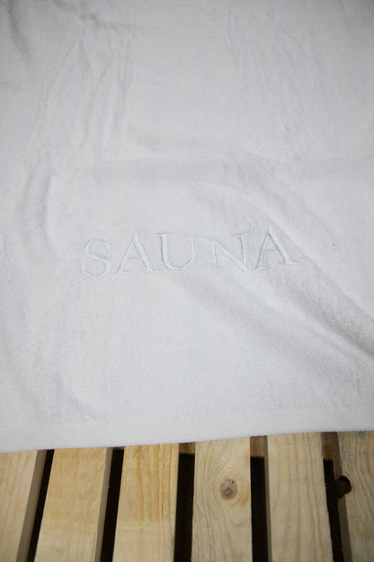 Auf einer Bank liegt ein Saunatuch mit aufgesticktem Text SAUNA von p.k. aus 100 Prozent Baumwolle in der Groesse 70x200cm. Farbe Weiss. Modell Weiss.