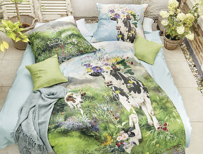 Auf einem Bett liegen mehrere Kopfkissen und eine Bettdecke bezogen mit Mako-Satin Bettwaesche aus 100 Prozent Baumwolle mit Muster aus Kuehen auf einer Wiese in der Farbe Gruen. Modell Almwiese.