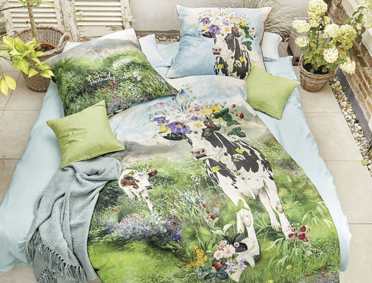 Auf einem Bett liegen mehrere Kopfkissen und eine Bettdecke bezogen mit Mako-Satin Bettwaesche aus 100 Prozent Baumwolle mit Muster aus Kuehen auf einer Wiese in der Farbe Gruen. Modell Almwiese.