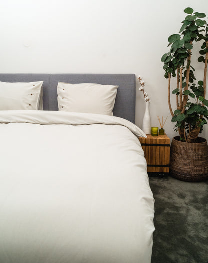 Auf einem Bett liegen mehrere Kopfkissen und eine Bettdecke bezogen mit Perkal-Baumwoll Bettwaesche aus 100 Prozent Baumwolle in der Farbe Silber. Modell Silbergrau.