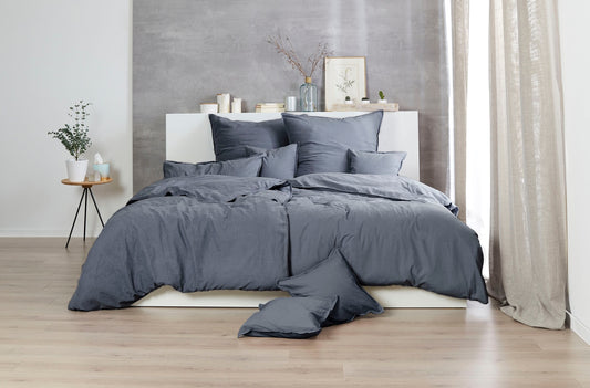 Auf einem Bett liegen mehrere Kopfkissen und eine Bettdecke bezogen mit Renforce Bettwaesche aus 100 Prozent Baumwolle in der Farbe Grau. Modell Stone-Washed Anthrazit 888.
