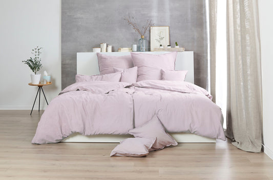 Auf einem Bett liegen mehrere Kopfkissen und eine Bettdecke bezogen mit Renforce Bettwaesche aus 100 Prozent Baumwolle in der Farbe Rosa. Modell Stone-Washed Rosenholz 450.