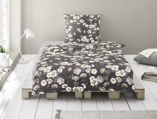 Auf einem Bett liegen mehrere Kopfkissen und eine Bettdecke bezogen mit Satin Bettwaesche aus 100 Prozent Baumwolle mit Muster aus Blumen in der Farbe Grau. Modell Blumen Grau.