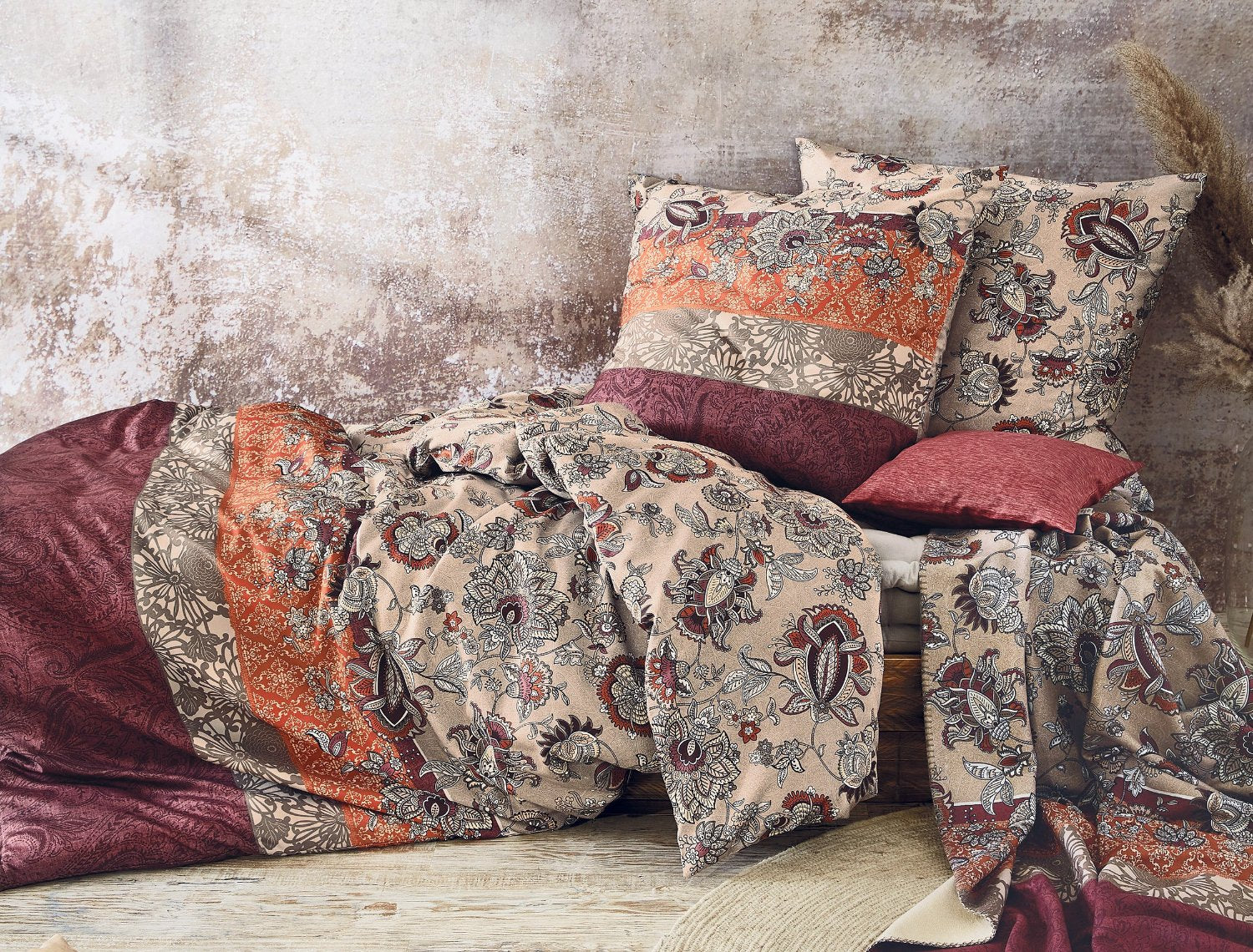 Auf einem Bett liegen mehrere Kopfkissen und eine Bettdecke bezogen mit Satin Bettwaesche aus 100 Prozent Baumwolle mit Muster aus Blumen in der Farbe Bordeaux. Modell EOS 01.
