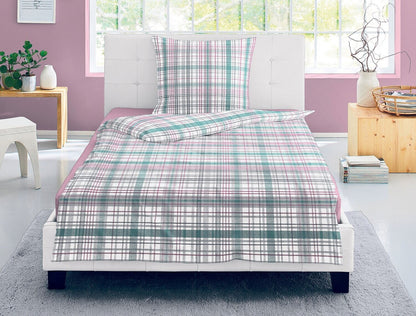 Auf einem Bett liegen mehrere Kopfkissen und eine Bettdecke bezogen mit Satin Bettwaesche aus 100 Prozent Baumwolle mit Muster aus Quadraten in der Farbe Rosa. Modell Karo Rosa Gruen.
