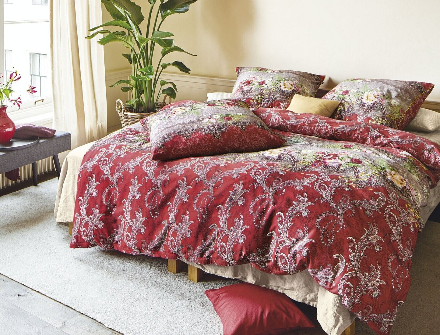 Auf einem Bett liegen mehrere Kopfkissen und eine Bettdecke bezogen mit Satin Bettwaesche aus 100 Prozent Baumwolle mit Muster aus Ornamenten in der Farbe Rot. Modell Oriental Rot.