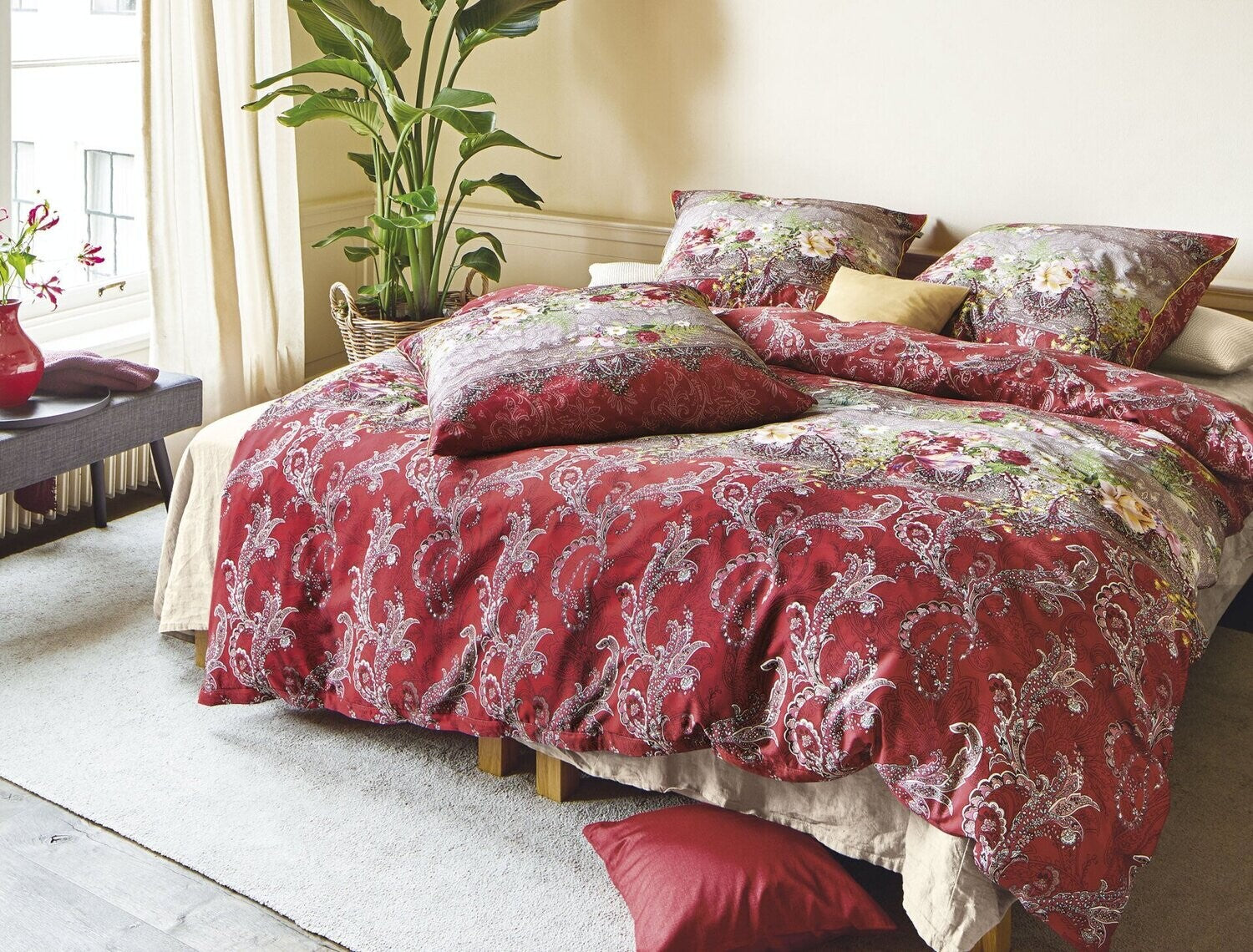 Auf einem Bett liegen mehrere Kopfkissen und eine Bettdecke bezogen mit Satin Bettwaesche aus 100 Prozent Baumwolle mit Muster aus Ornamenten in der Farbe Rot. Modell Oriental Rot.