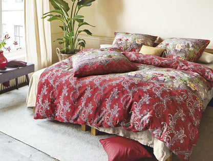 Auf einem Bett liegen mehrere Kopfkissen und eine Bettdecke bezogen mit Satin Bettwaesche aus 100 Prozent Baumwolle mit Muster aus Ornamenten in der Farbe Rot. Modell Oriental Rot.