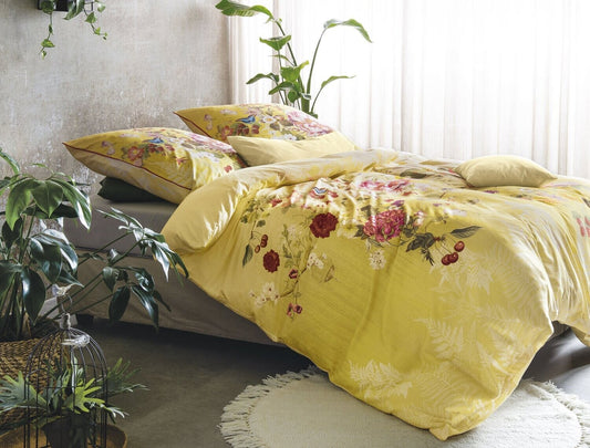 Auf einem Bett liegen mehrere Kopfkissen und eine Bettdecke bezogen mit Satin Bettwaesche aus 100 Prozent Baumwolle mit Muster aus Blumen in der Farbe Gelb. Modell Rose Gelb.