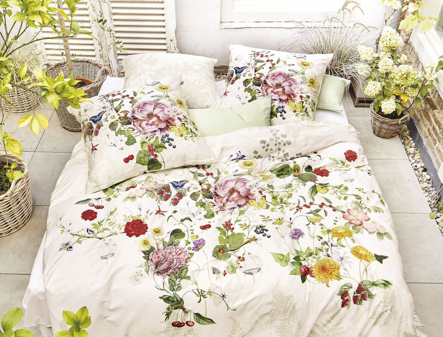 Auf einem Bett liegen mehrere Kopfkissen und eine Bettdecke bezogen mit Satin Bettwaesche aus 100 Prozent Baumwolle mit Muster aus Blumen in der Farbe Weiss. Modell Rose Weiss.