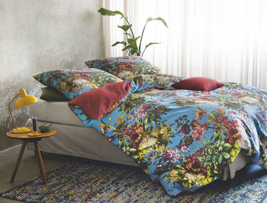 Auf einem Bett liegen mehrere Kopfkissen und eine Bettdecke bezogen mit Satin Bettwaesche aus 100 Prozent Baumwolle mit Muster aus Blumen in der Farbe Blau. Modell Sommer Blau.