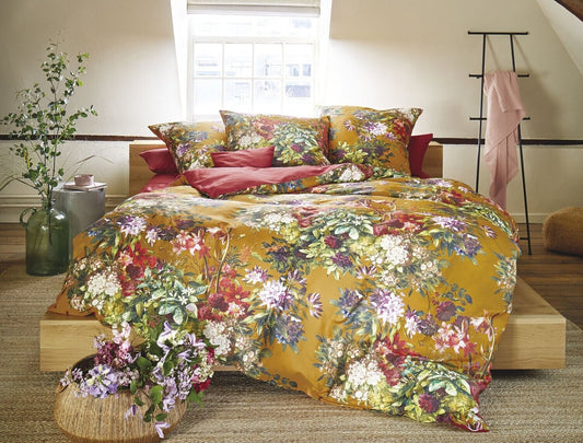Auf einem Bett liegen mehrere Kopfkissen und eine Bettdecke bezogen mit Satin Bettwaesche aus 100 Prozent Baumwolle mit Muster aus Blumen in der Farbe Gelb. Modell Sommer Gelb.