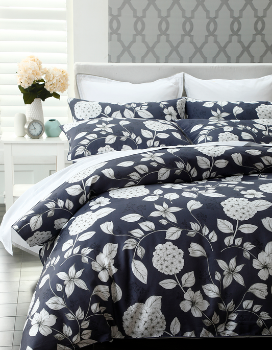 Auf einem Bett liegen mehrere Kopfkissen und eine Bettdecke bezogen mit Satin Bettwaesche aus 100 Prozent Baumwolle mit Muster aus Blumen in der Farbe Blau. Modell Hydrangea.